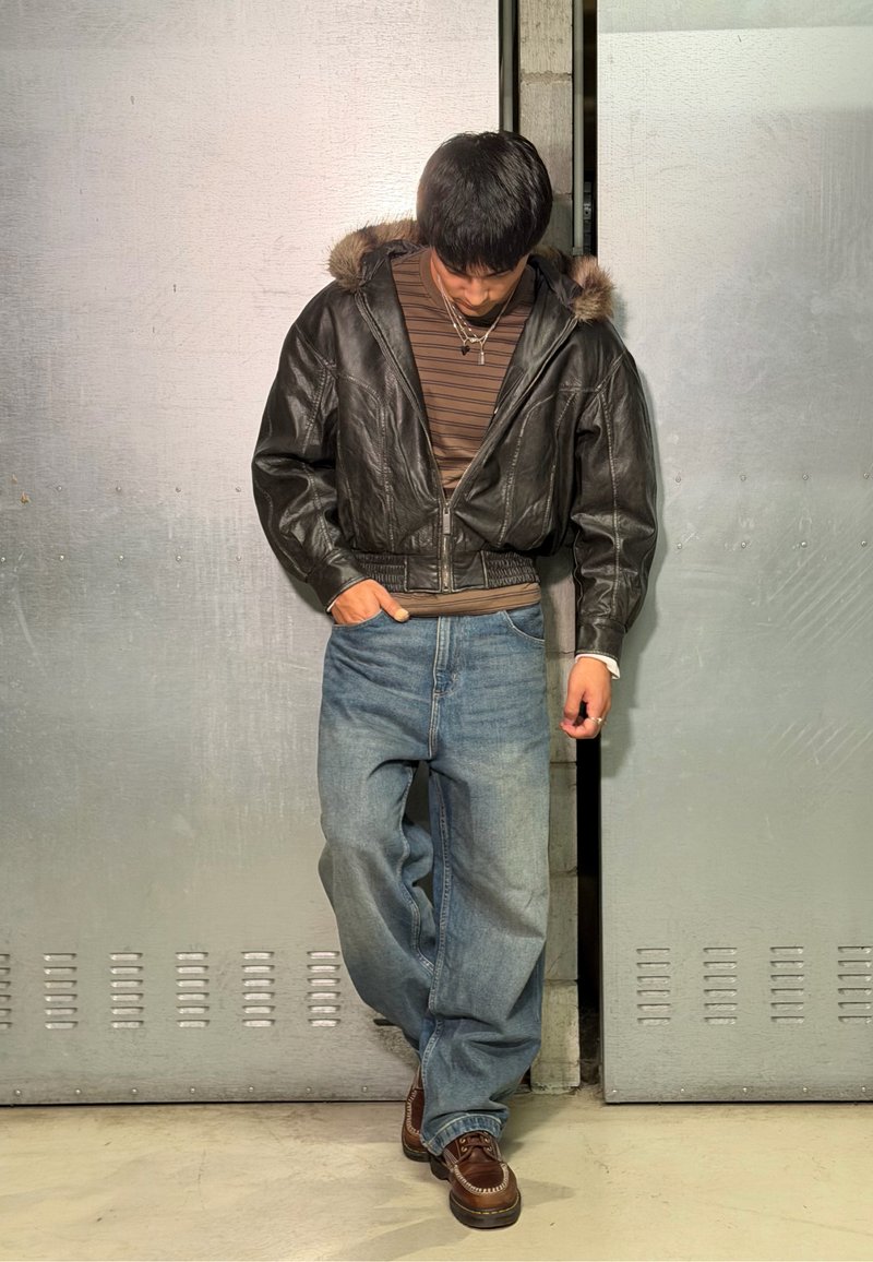 Jeune homme portant une chemise marron rayée, une veste en cuir noire, un jean bleu ample et des chaussures brunes, se tient contre des portes métalliques en regardant vers le bas.