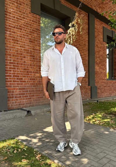 Un hombre está de pie contra una pared de ladrillo, vistiendo una camisa blanca a rayas, pantalones beige claros y zapatillas grises, sosteniendo una tableta oscura o una carpeta.