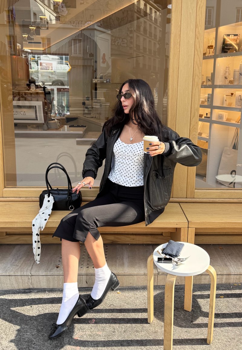 Veste bomber en cuir noir sur un haut à pois blancs, pantalon noir court et chaussures noires avec des chaussettes blanches. Les accessoires incluent un sac à main noir et un café.