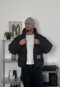Must kärgaga puffer-jakk, mis on varustatud lukuga eest, kapuutsiga, bordüüriga "LIVE FAST" logo ja kahe küljetaskuga. Kombineeritud halli beanie'ga.