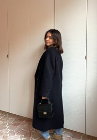 Femme aux cheveux mi-longs portant un long manteau noir, un jean bleu clair et des bottes beiges, tenant un petit sac à main noir à l'intérieur.