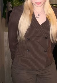 Blonde person wearing a brown pinstripe double-breasted blazer and matching trousers, standing with hands behind their back.