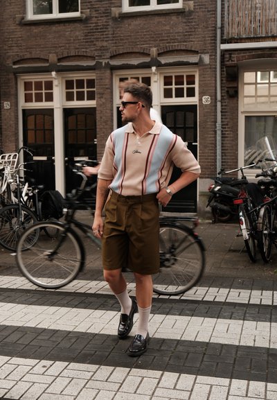 Polo de punto en beige con rayas azul claro y rojas; pantalones cortos marrones de corte ajustado; mocasines negros; calcetines blancos; fondo urbano.