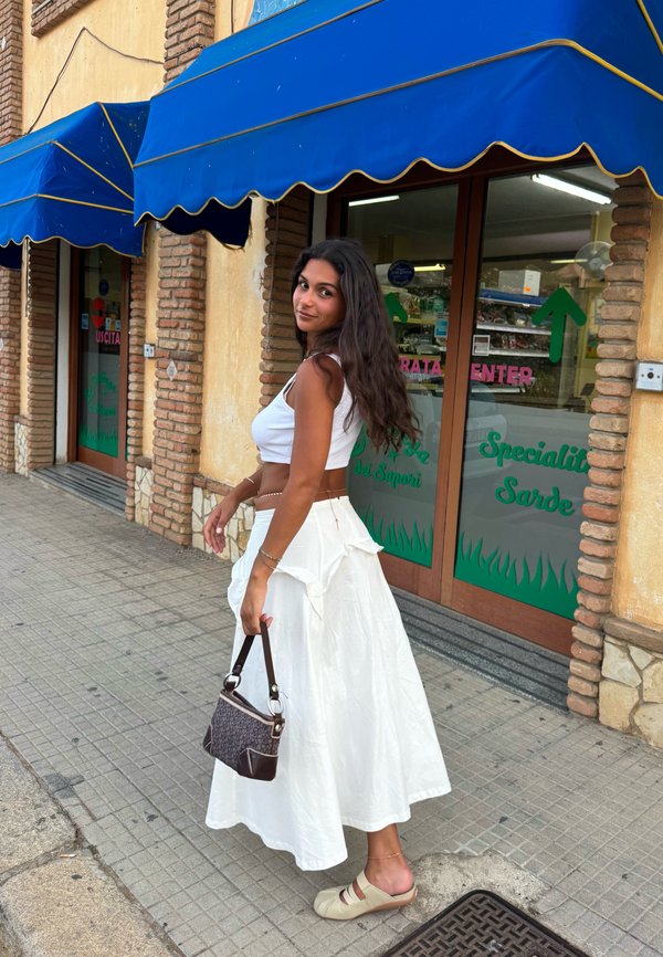 Femme en jupe et haut blancs tenant un sac à main, debout sur le trottoir devant un bâtiment en briques avec des auvents bleus et des portes en verre.