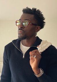 Mężczyzna o kręconych włosach, noszący przyciemniane okulary przeciwsłoneczne, czarny sweter zapinany na zamek i srebrną biżuterię, trzymający białą teksturowaną ściereczkę na ramieniu.