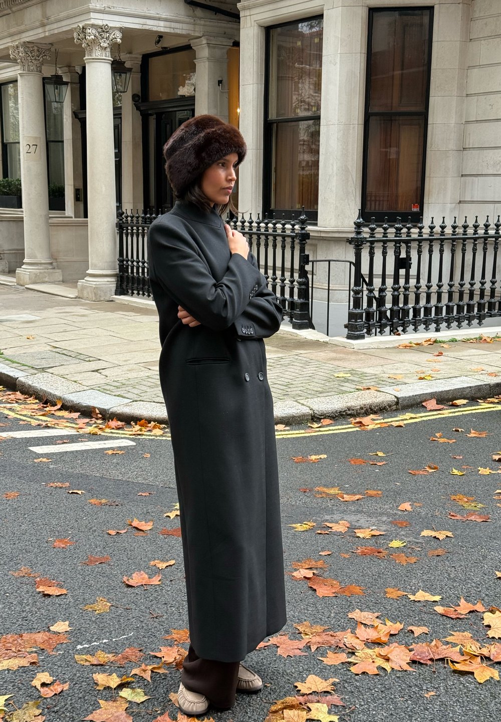 Hosszú fekete kabát kettős gombokkal és magas gallérral, barna szőrmés sapkával párosítva, háttérben falevelekkel borított utca.