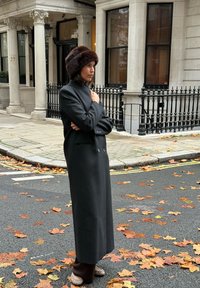 Hosszú fekete kabát kettős gombokkal és magas gallérral, barna szőrmés sapkával párosítva, háttérben falevelekkel borított utca.