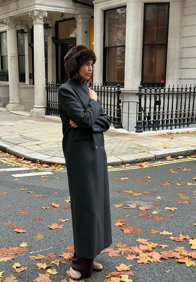 Abrigo negro largo con botones dobles y cuello alto, combinado con un sombrero de piel marrón, ambientado en una calle con hojas caídas.