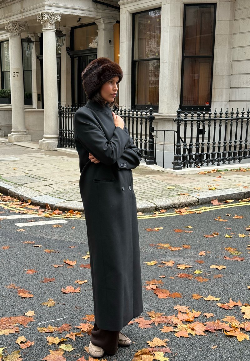 Langer schwarzer Mantel mit doppelter Knopfleiste und hohem Kragen, kombiniert mit einem braunen Pelzhut, vor einer Straße mit herabgefallenen Blättern.