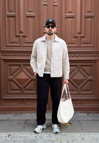 Chemise boutonnière crème, pull beige, pantalon noir, baskets argentées et sac fourre-tout blanc devant un mur avec motif géométrique marron.