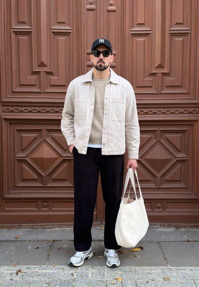 Camisa de botones color crema, suéter beige, pantalones negros, zapatillas plateadas y bolso blanco contra una pared marrón con patrón geométrico.
