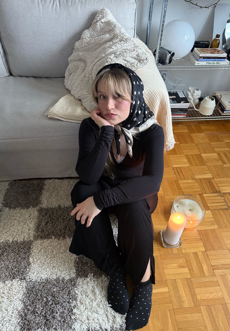 Jeune femme en tenue noire et foulard à pois assise par terre à côté d'une bougie allumée, d'un tapis doux et d'un canapé gris avec des coussins texturés.