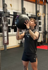 Homme tatoué tenant un ballon médicinal noir au-dessus de la tête dans une salle de sport avec des anneaux d'entraînement et des banderoles "ÉQUILIBRE" en arrière-plan.
