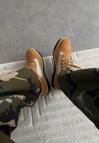 Bottes de randonnée marron et beige avec des lacets jaunes et des semelles en caoutchouc noir, portées avec un pantalon à motif camouflage, reposant sur un tapis blanc texturé.