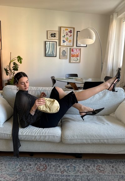Mujer recostada en un sofá gris claro con top negro de lunares, pantalones negros y tacones negros, sosteniendo un pequeño bolso beige en una sala de estar moderna.