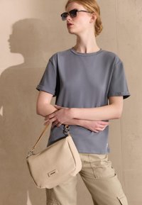 Graues kurzärmliges T-Shirt kombiniert mit khakifarbenen Cargo-Hosen. Eine strukturierte beigen Schultertasche mit kurzem Tragegurt und silbernen Beschlägen wird gehalten.