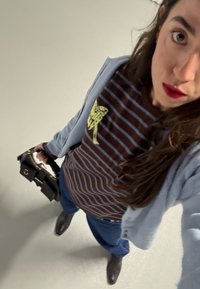 Femme tenant un sac à main noir, portant une chemise rayée marron et bleue avec un motif jaune, un cardigan bleu clair, un jean bleu et des bottes marron.