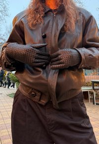 Άτομο που φοράει μαύρα πλεκτά δερμάτινα γάντια και μαύρο δερμάτινο μπουφάν σε εξωτερικό χώρο, με δέντρα και ανθρώπους στο φόντο.