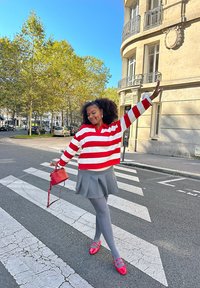 Rödvit randig tröja, grå veckad kjol, grå strumpbyxor och röda skor. Håller en liten röd handväska i en övergångsställe. Klar himmel.