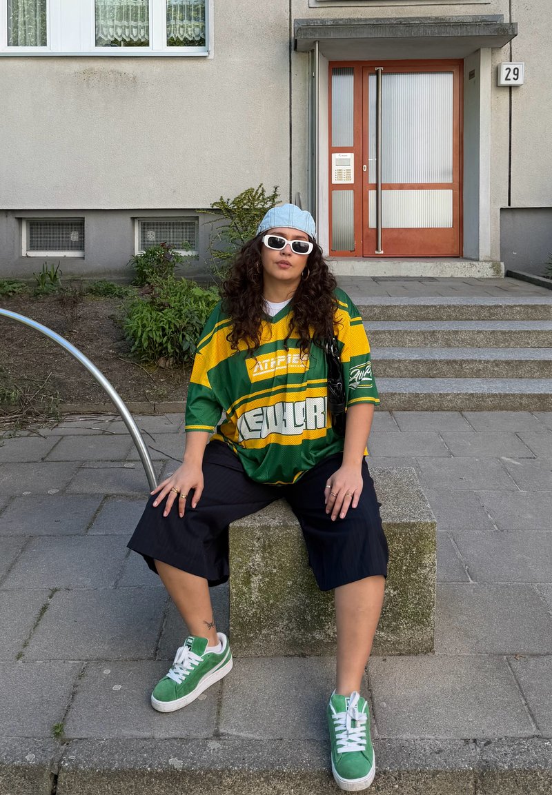 Personne aux cheveux bouclés portant des lunettes de soleil, une casquette, une chemise oversized verte et jaune, et des baskets vertes, assise sur un bloc de pierre à l'extérieur d'un bâtiment.