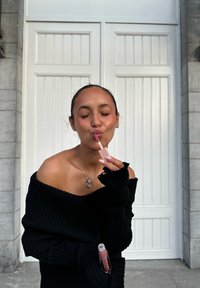 Zwarte geribbelde off-shoulder trui, lichtroze lipgloss-applicator in de hand, zilveren sieraden, subtiele blush, textuur achtergrond met witte deuren.