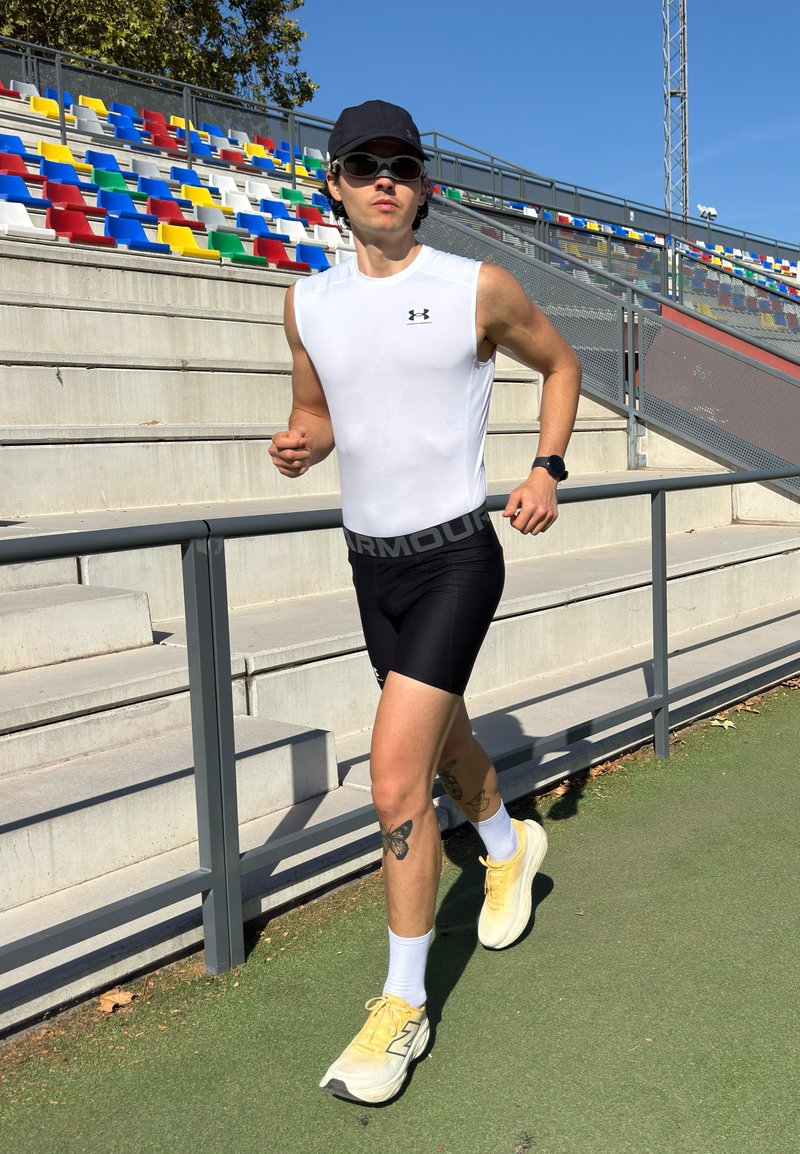 Jeune homme faisant du jogging sur une piste verte à côté de gradins vides d'un stade avec des sièges colorés, portant une casquette noire, des lunettes de soleil et des vêtements de sport.