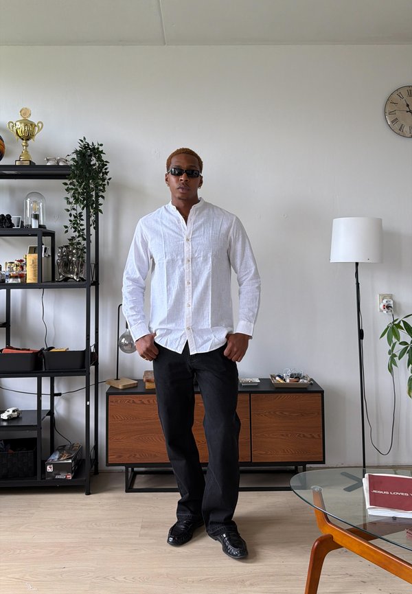 Homme portant des lunettes de soleil, une chemise blanche boutonnée et un pantalon noir se tient dans le salon avec des meubles en bois, une plante et une horloge au mur.