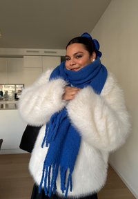 Fehér műszőrme kabát széles ujjakkal, kék texturált sállal körbetekerve, és egy fekete táska. A háttér egyszerű konyhai elemeket ábrázol.