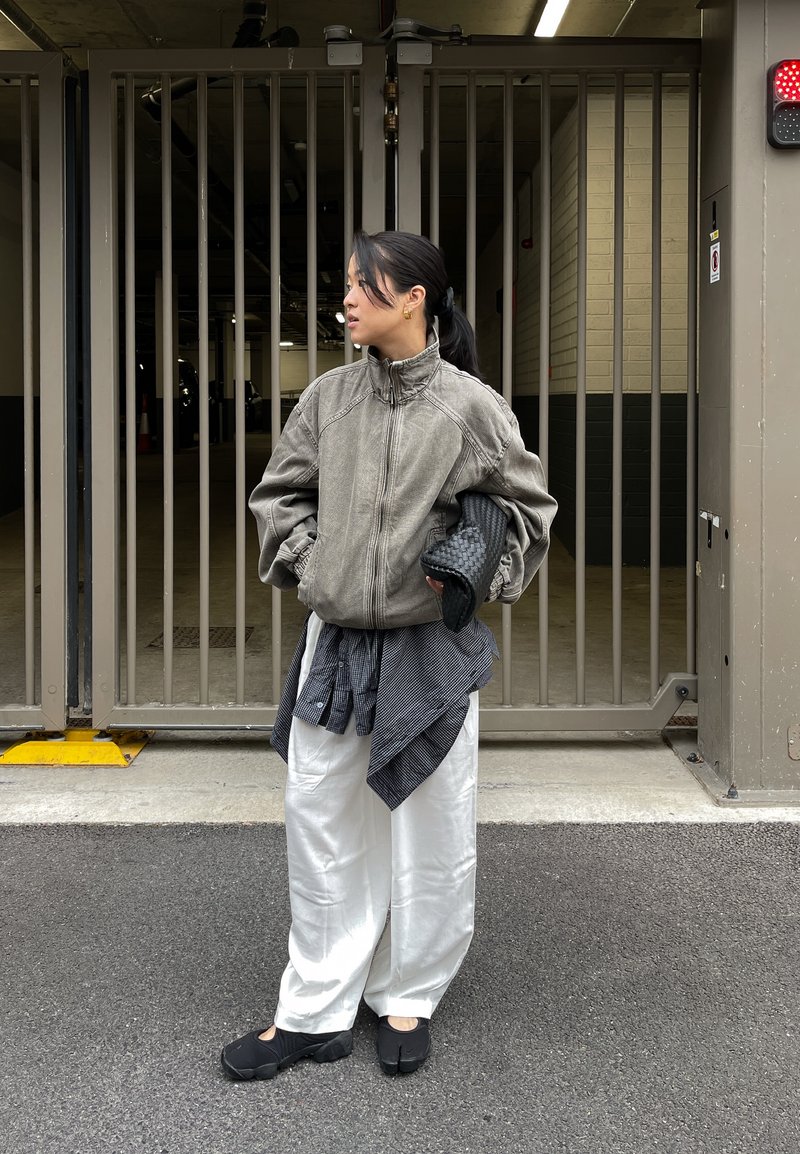 Veste grise surdimensionnée avec un col montant, pantalon blanc à jambes larges, et chaussures noires. Accessoires comprenant un sac noir texturé et une chemise à carreaux nouée à la taille.