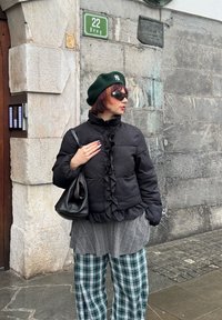 Femme portant un béret vert, des lunettes de soleil à motif d'étoiles, une veste noire à volants, une chemise grise et un pantalon à carreaux verts, debout contre un mur en pierre, tenant un sac noir.