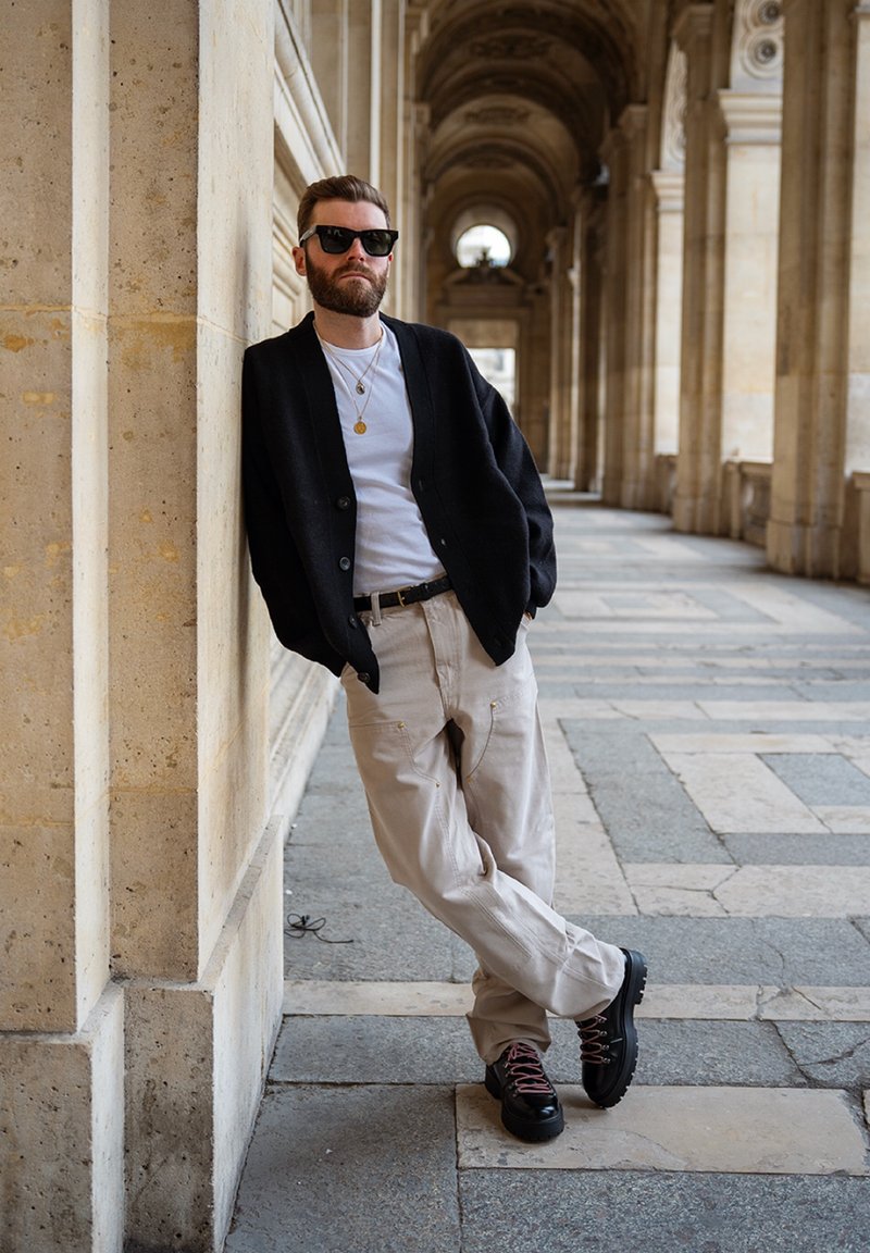 Homme portant des lunettes de soleil, cardigan noir, chemise blanche, pantalon beige et bottes noires, appuyé contre un mur en pierre dans un couloir extérieur voûté.