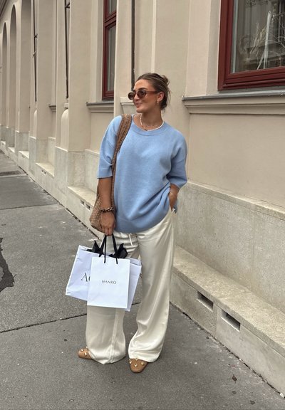 Suéter de punto azul claro, pantalones de pierna ancha crema ajustados, llevando bolsas de compras blancas, usando gafas de sol marrón oversized y mules beige con adornos.