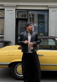 Svart lærbomberjakke, grå henley-skjorte, løse svarte bukser, holder en drikke, står ved siden av en gul klassisk bil.