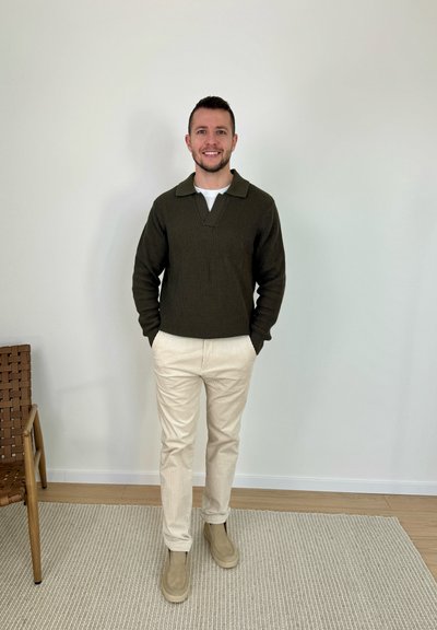 Hombre sonriendo, con suéter verde oscuro, camiseta blanca, pantalones beige y botas beige, de pie en el interior sobre una alfombra junto a una silla tejida.