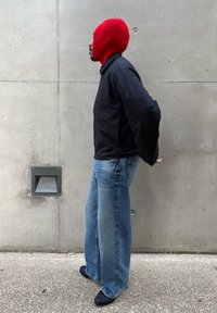 Fekete dzseki, világoskék, bőszárú farmernadrág és élénkpiros kötött balaklava. A háttér sima, szürke betonfal.