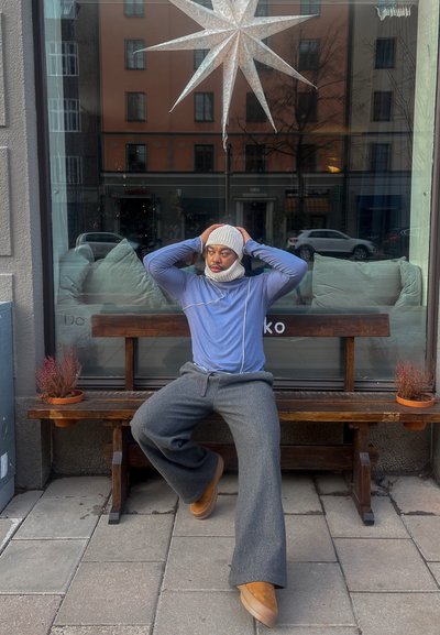 Persona sentada en un banco de madera, usando un gorro gris, una camisa de manga larga azul con detalles blancos, pantalones anchos grises y calzado beige.