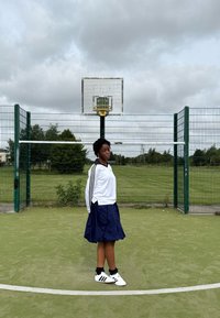 Atleta con chaqueta de deporte blanca de Adidas, falda azul marino y zapatillas blancas con rayas negras, de pie en una cancha verde con un aro de baloncesto.