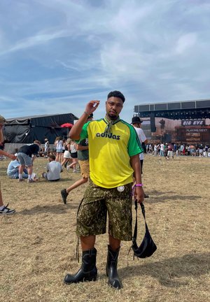 Grün und gelbe Adidas-Jersey, CAMO-Shorts und schwarze kniehohe Stiefel. Hält eine Umhängetasche und steht auf Gras bei einem Festival.