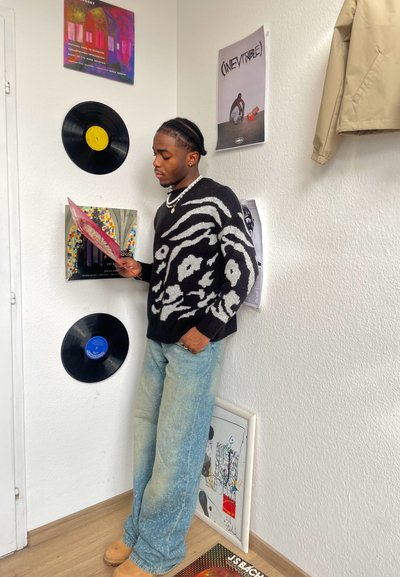 Persona vestida con un suéter negro y gris con patrón, jeans claros desgastados y zapatos color tostado, sosteniendo un disco de vinilo entre arte en la pared y discos.
