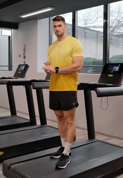 Hombre con camiseta amarilla y pantalones cortos negros de pie en una cinta de correr en un gimnasio, con grandes ventanas y múltiples cintas de correr visibles.