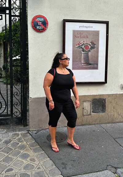 Top negro ajustado y pantalones recortados, chanclas rojas, gafas de sol; de pie junto a una pared con un cartel y un letrero redondo que dice "JOUR ET NUIT."