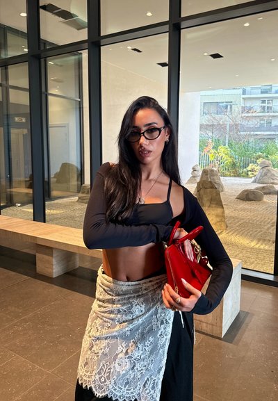 Mujer con cabello largo y oscuro, vistiendo un top corto negro, falda de encaje y gafas, sosteniendo un bolso rojo dentro de un edificio moderno.