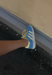 Perna estendida, vestindo sapatilhas Adidas azuis e amarelas, apoiada contra um meio-fio cinza e uma parede bege, sobre um chão de asfalto.