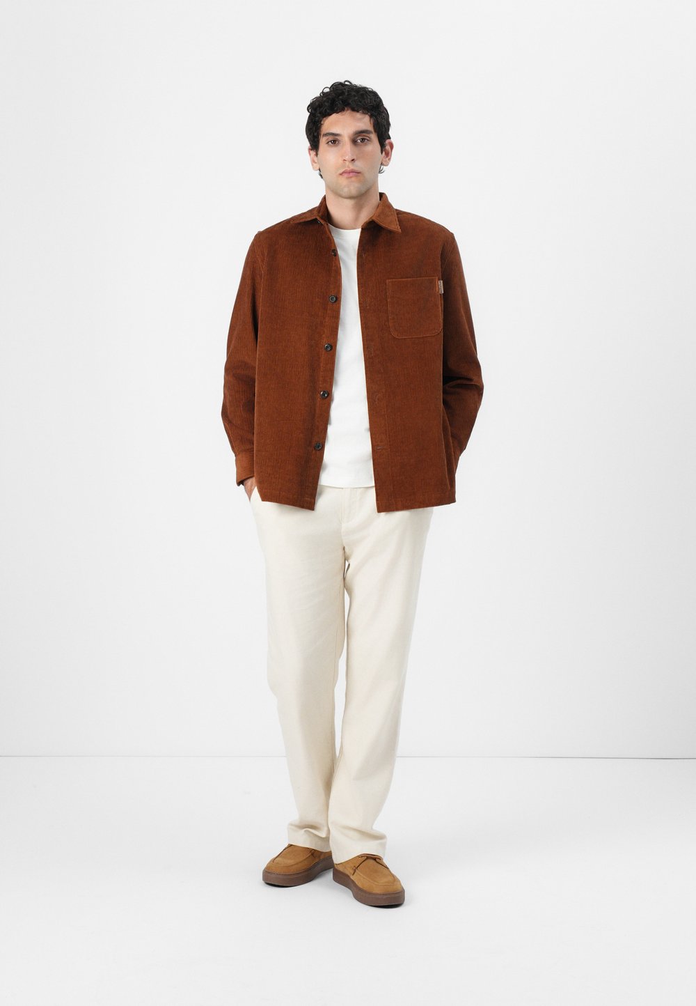 Marrón chaleco de pana con un solo bolsillo en el pecho, combinado con pantalones crema y zapatos marrones. El modelo está de pie frente a un fondo liso.