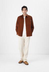 Marrón chaleco de pana con un solo bolsillo en el pecho, combinado con pantalones crema y zapatos marrones. El modelo está de pie frente a un fondo liso.