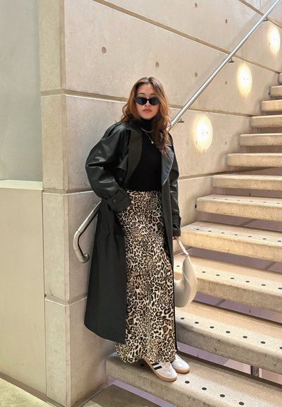Abrigo de cuero negro, cuello alto negro, falda de leopardo, zapatillas blancas y gafas de sol, posando contra una escalera con paredes texturizadas.