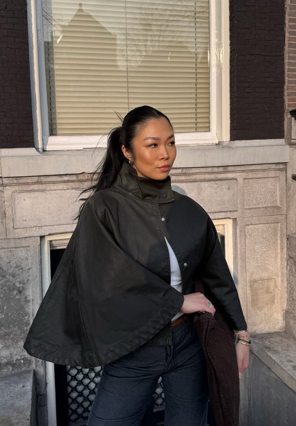 Jonge vrouw met lang donker haar in een hoge paardenstaart draagt een zwart cape-jasje, staand buiten naast een stenen gebouw met een raam en jaloezieën.