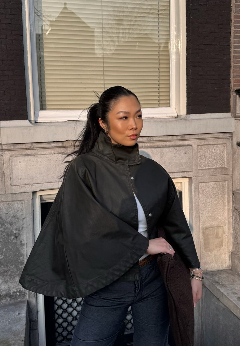 Jeune femme aux longs cheveux noirs attachés en queue de cheval haute, portant une veste cape noire, debout à l'extérieur près d'un bâtiment en pierre avec une fenêtre et des stores.