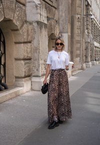 T-shirt blanc, jupe maxi à imprimé léopard, lunettes de soleil noires. Tenant une tasse de café, portant des chaussures compensées noires, debout dans une rue.