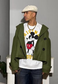 Vihreä lyhyt takki, jossa isot mustat napit, valkoisen graafisen Mickey Mouse -kuvioidun T-paidan päällä. Tumma siniset farkut ja valkoinen lippalakki.